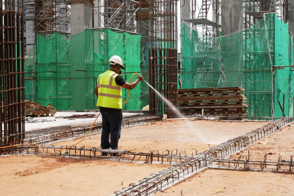 Worker in a hard hat and hi-vis vest spraying the ground between rebar on a construction site before the slab is poured